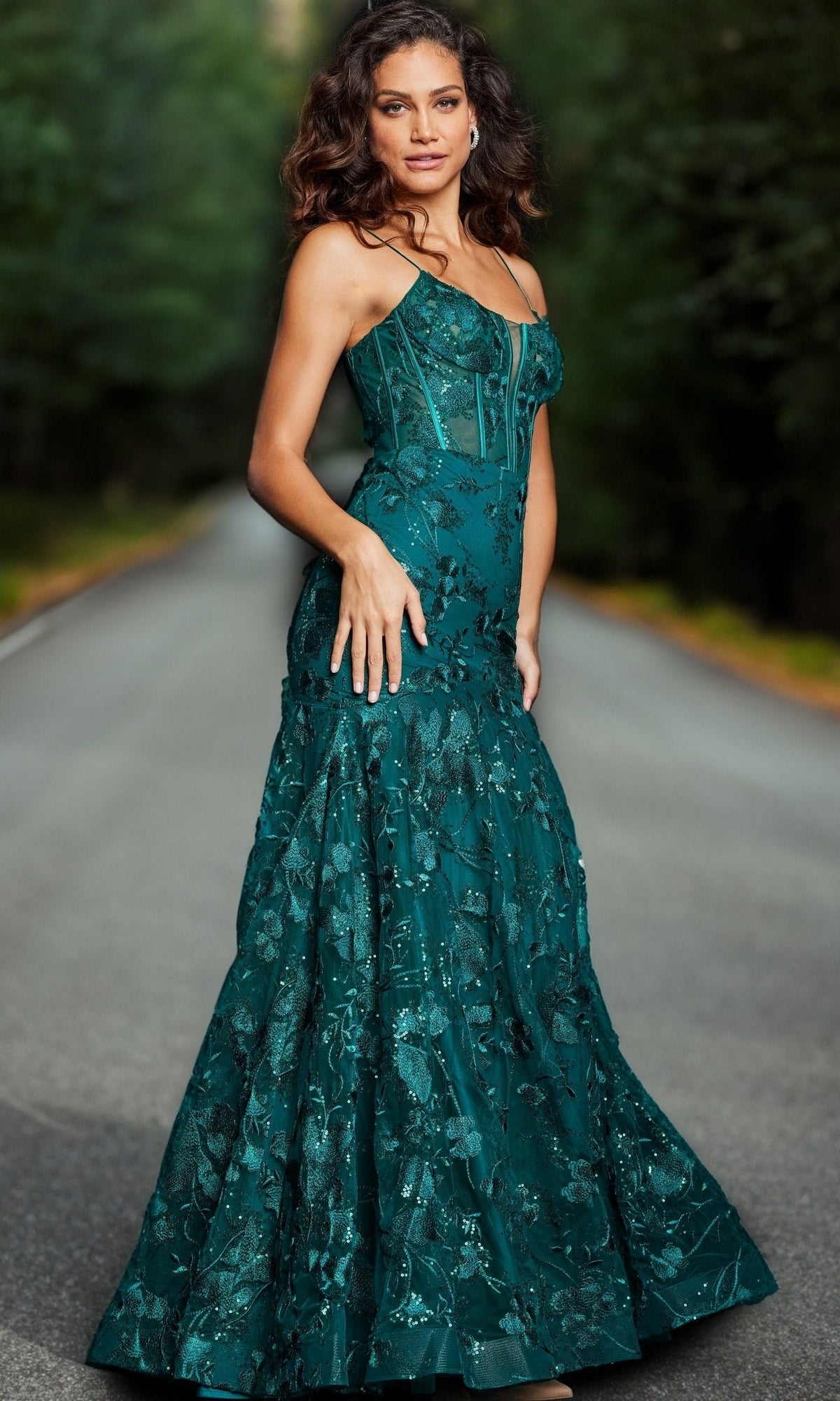 A woman in the Jovani 38004 Long Corset Mermaid Prom Dress, featuring emerald green embroidered lace, stands on an empty tree-lined road, looking at the camera.